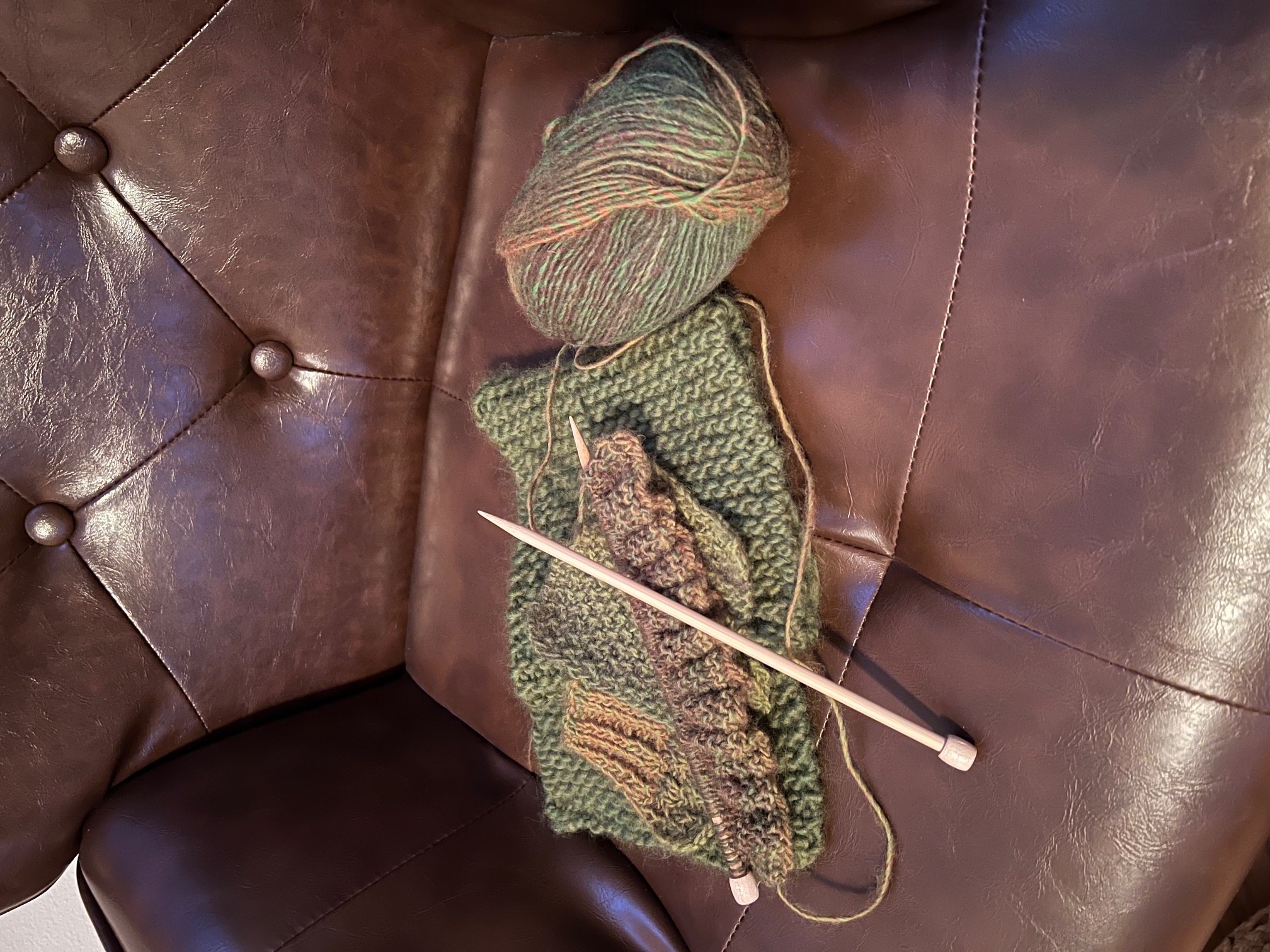 A photo of a faux leather chair with green yarn and a few practice pieces of knitting along with a work in progress with bamboo knitting needles.